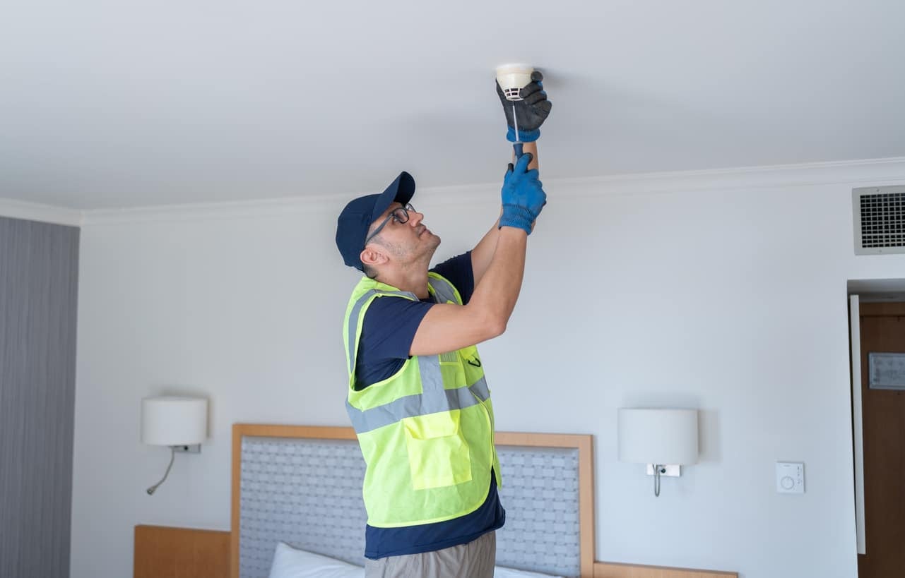 Worker Installing Smoke Detector At Hotel Room Ceiling